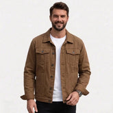 A smiling man wearing a coffee button-up trucker jacket over a white t-shirt and black pants, photographed against a clean white studio background.