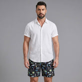 Man wearing a white linen rayon button up shirt and black palm tree print swim shorts front view on plain background