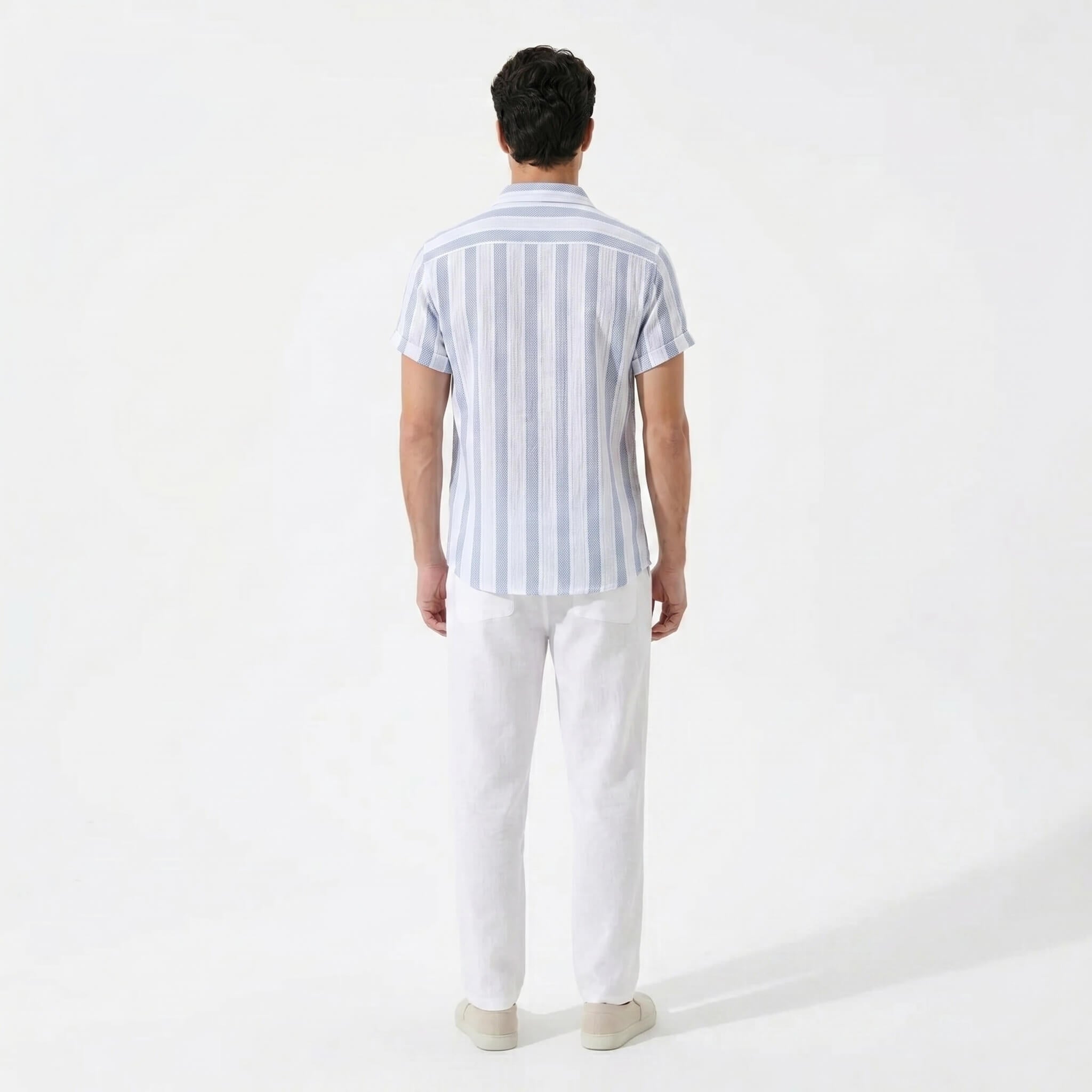 Back view of man wearing a light blue striped shirt and white pants on a white background
