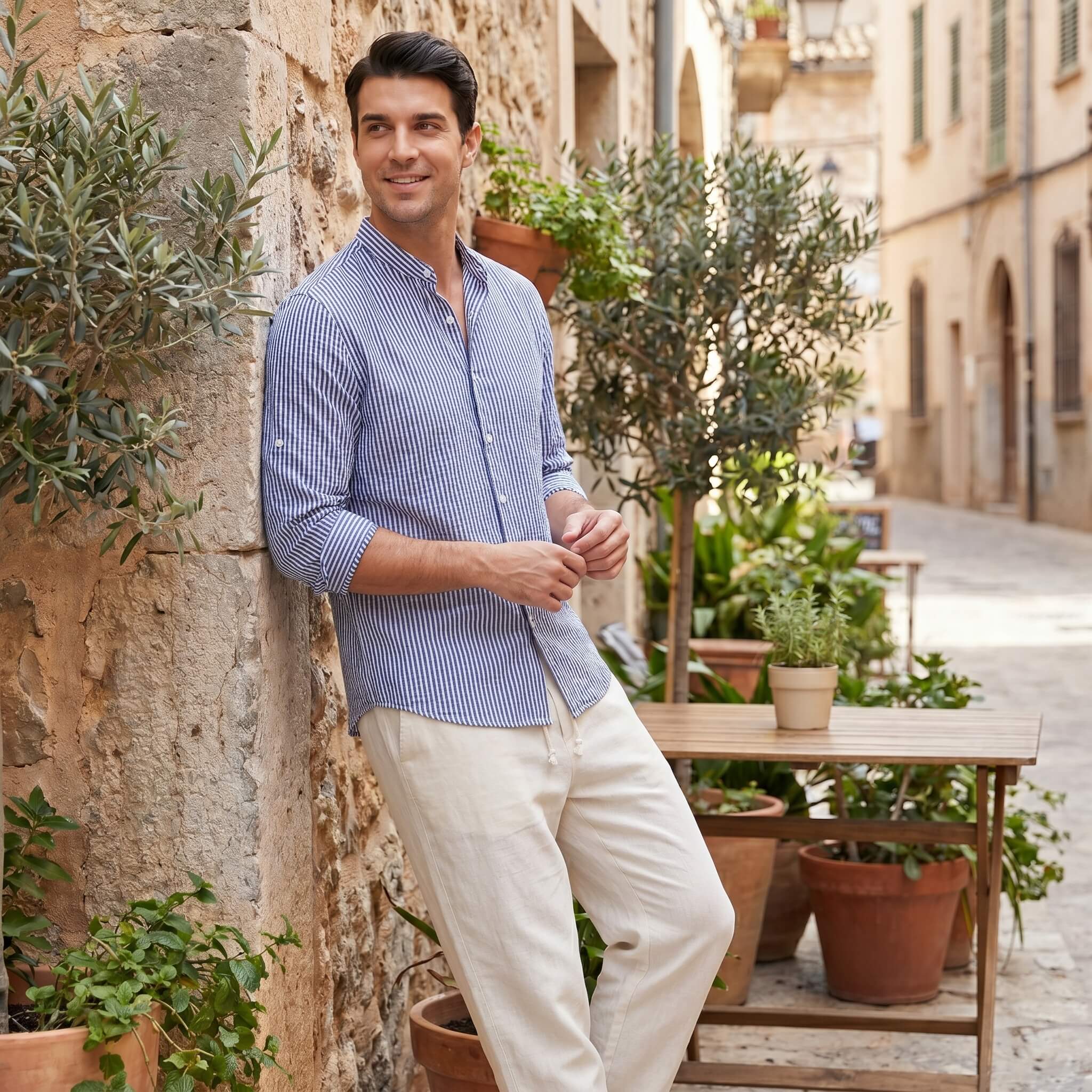 Man in a blue striped shirt and beige pants standing in an outdoor setting with plants and a table.