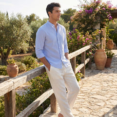 Man in a light blue shirt and beige pants standing on a stone path with greenery and flowers in the background.