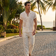 Man in a patterned shirt and beige pants walking on a paved path with palm trees and ocean in the background.