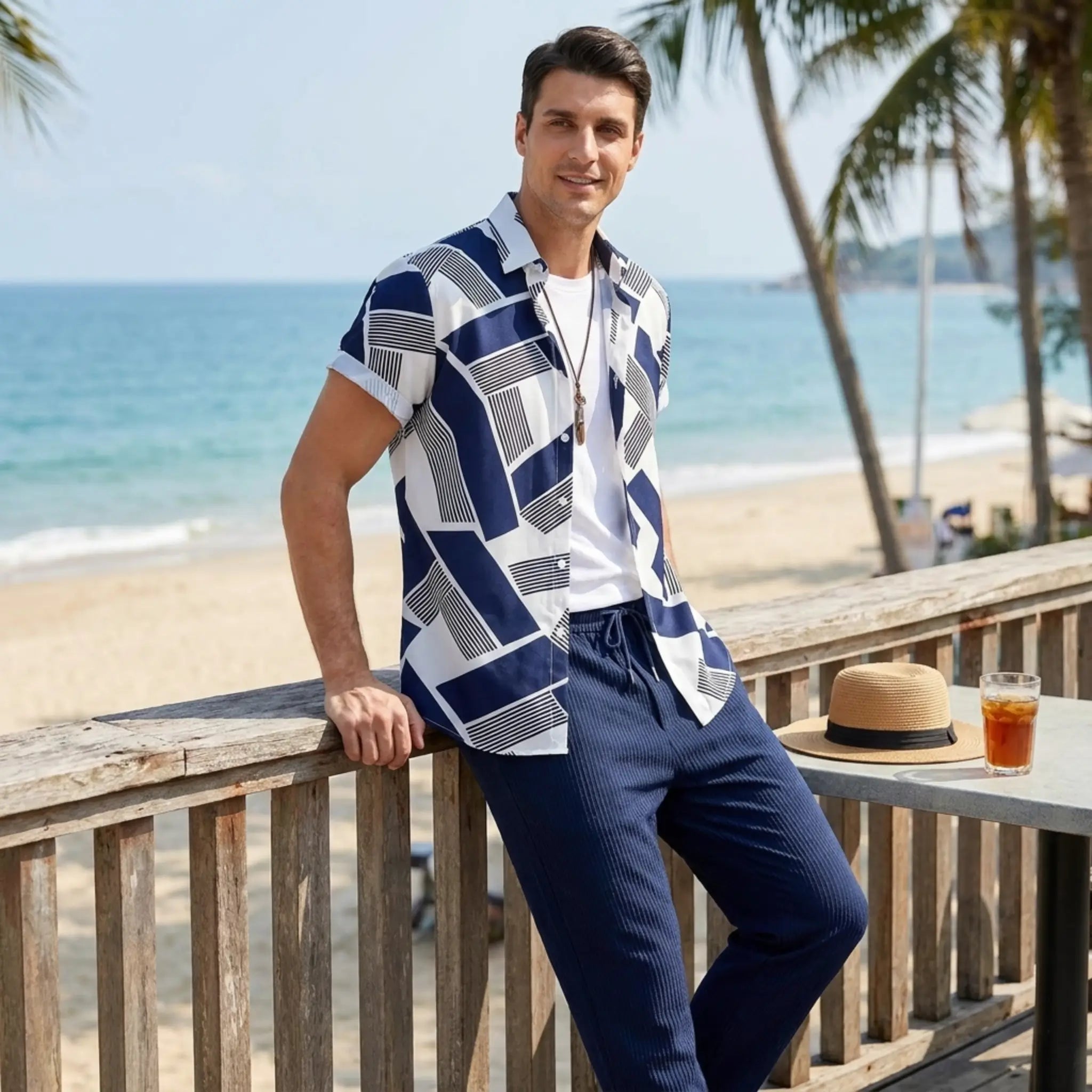 Man in a patterned shirt and navy pants leaning on a wooden railing by the beach.