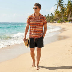Man in a patterned shirt and shorts walking on a beach with palm trees in the background