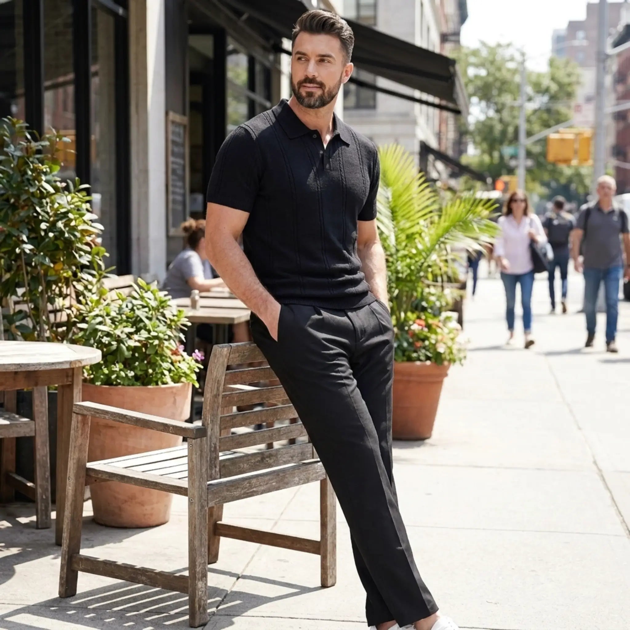 Man in black outfit leaning against a wooden chair on a city street.