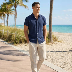 Man in navy shirt and light pants standing on a beach boardwalk with palm trees and ocean in the background.