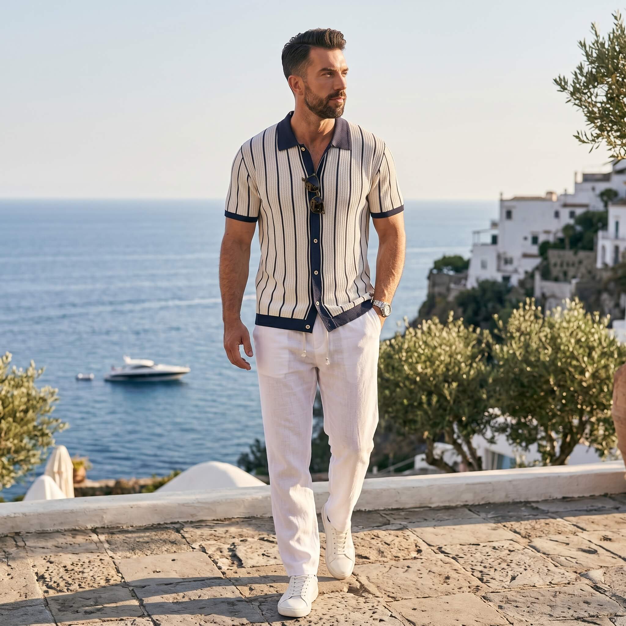 Man in striped shirt and white pants standing on a scenic coastal path with ocean and buildings in the background.