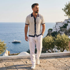 Man in striped shirt and white pants standing on a scenic coastal path with ocean and buildings in the background.