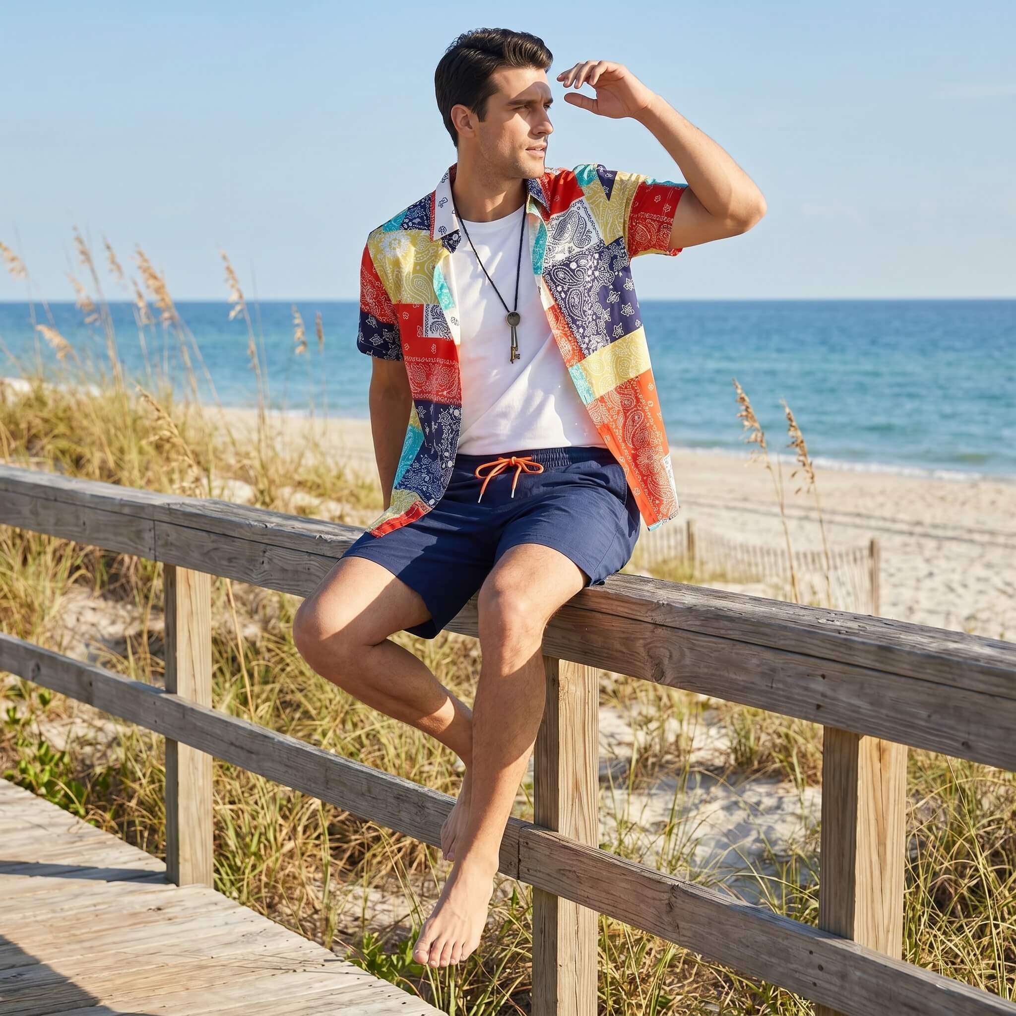 Man sitting on a wooden railing at the beach wearing a colorful shirt and navy shorts.