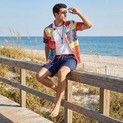 Man sitting on a wooden railing at the beach wearing a colorful shirt and navy shorts.