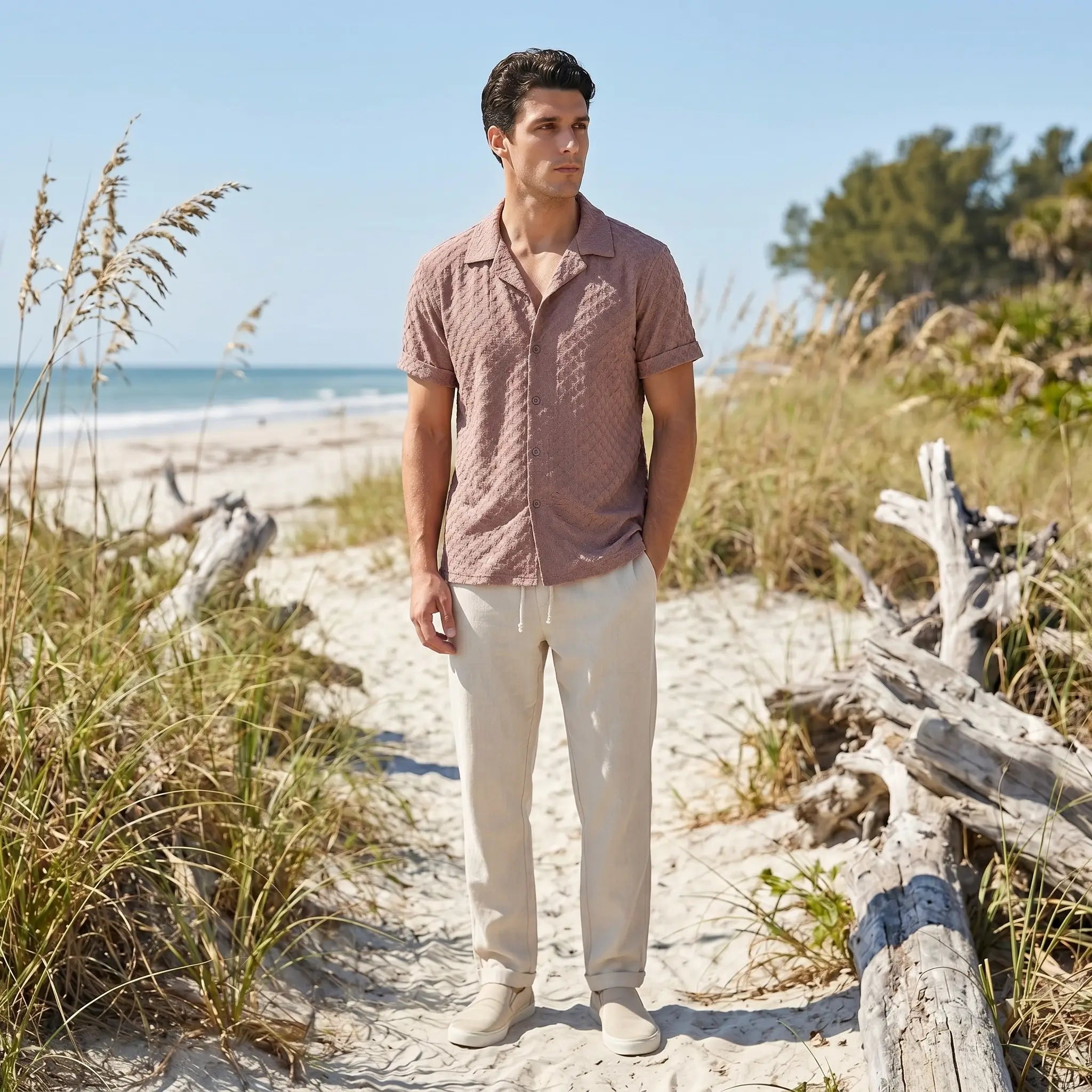 Man standing on a sandy path with beach grass and driftwood, wearing a patterned tan shirt and light pants.