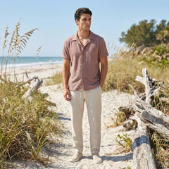 Man standing on a sandy path with beach grass and driftwood, wearing a patterned tan shirt and light pants.