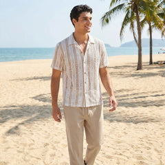 Man walking on a beach wearing a white patterned shirt and beige pants.
