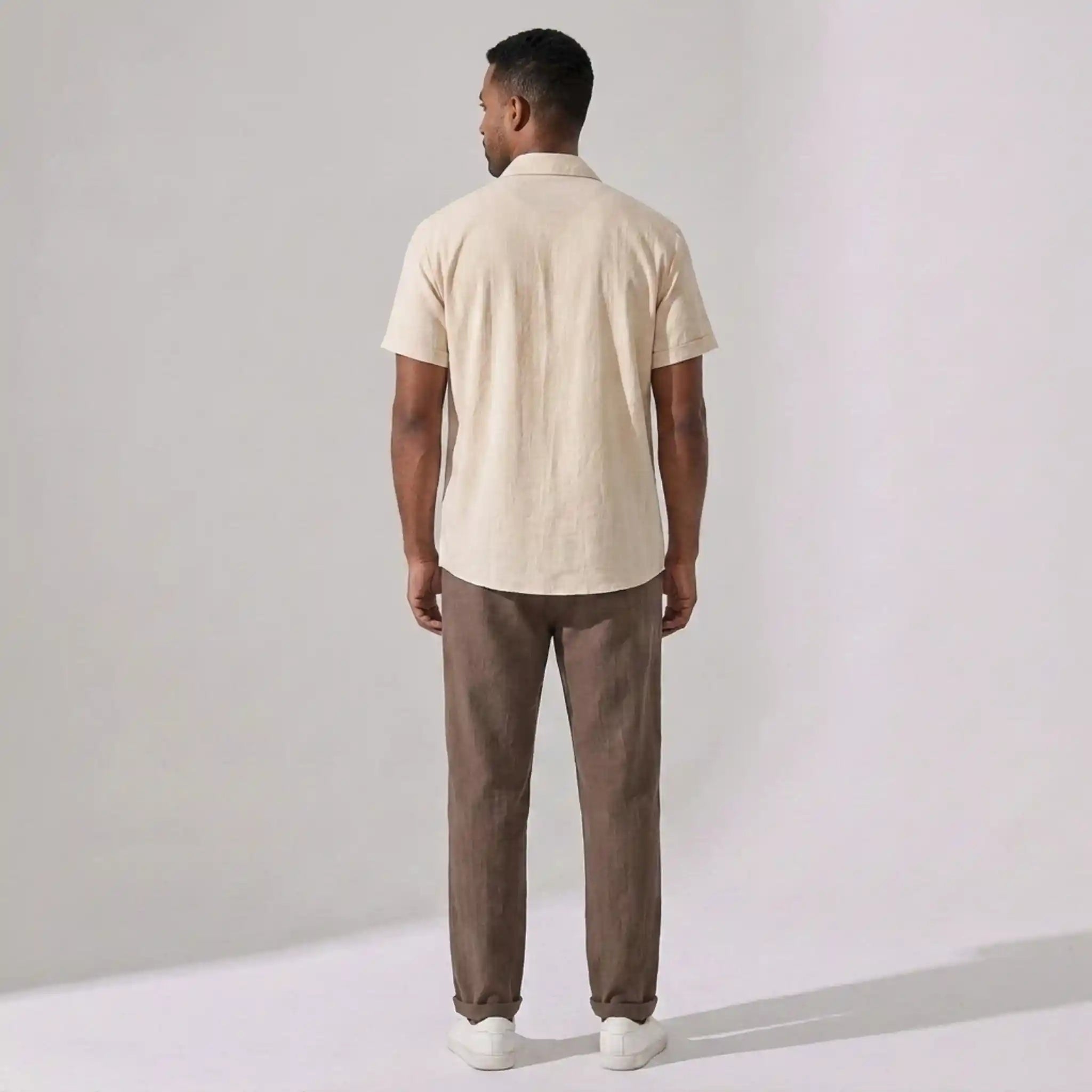 Man wearing a beige shirt and brown pants standing against a plain background back view