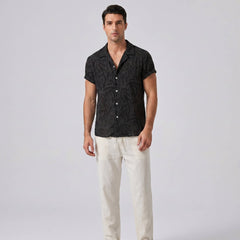 Man wearing a black patterned shirt and white pants on a light gray background
