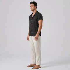 Man wearing a black patterned shirt and white pants on a plain background