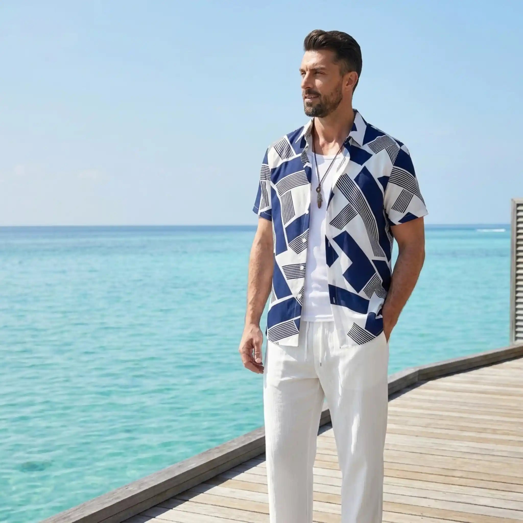 Man wearing a blue and white patterned shirt and white pants standing on a wooden deck by the ocean.