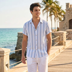 Man wearing a blue and white striped shirt and white pants standing on a coastal path with palm trees and ocean in the background.
