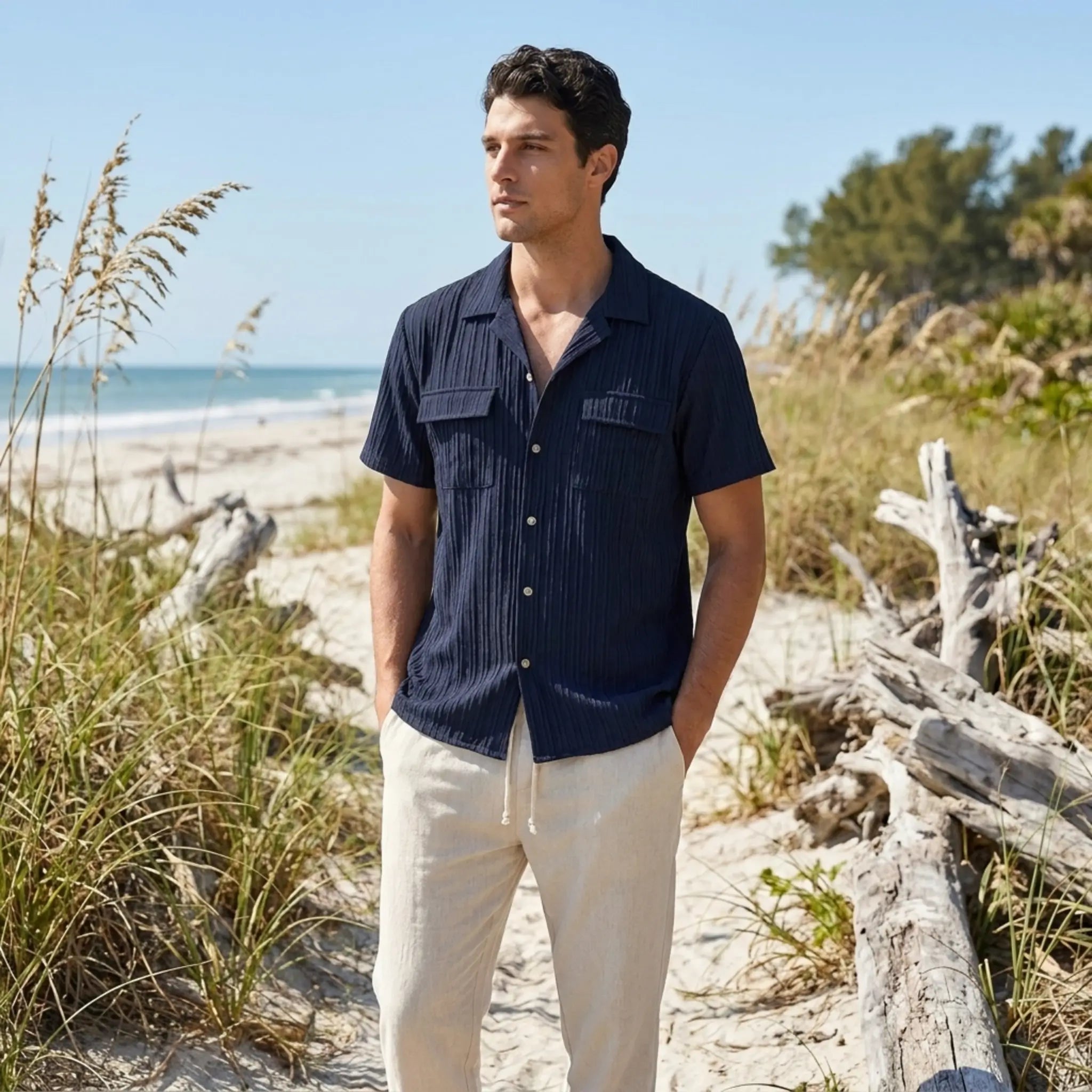 Man wearing a navy shirt and white pants standing on a beach with ocean and sky in the background