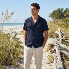 Man wearing a navy shirt and white pants standing on a beach with ocean and sky in the background