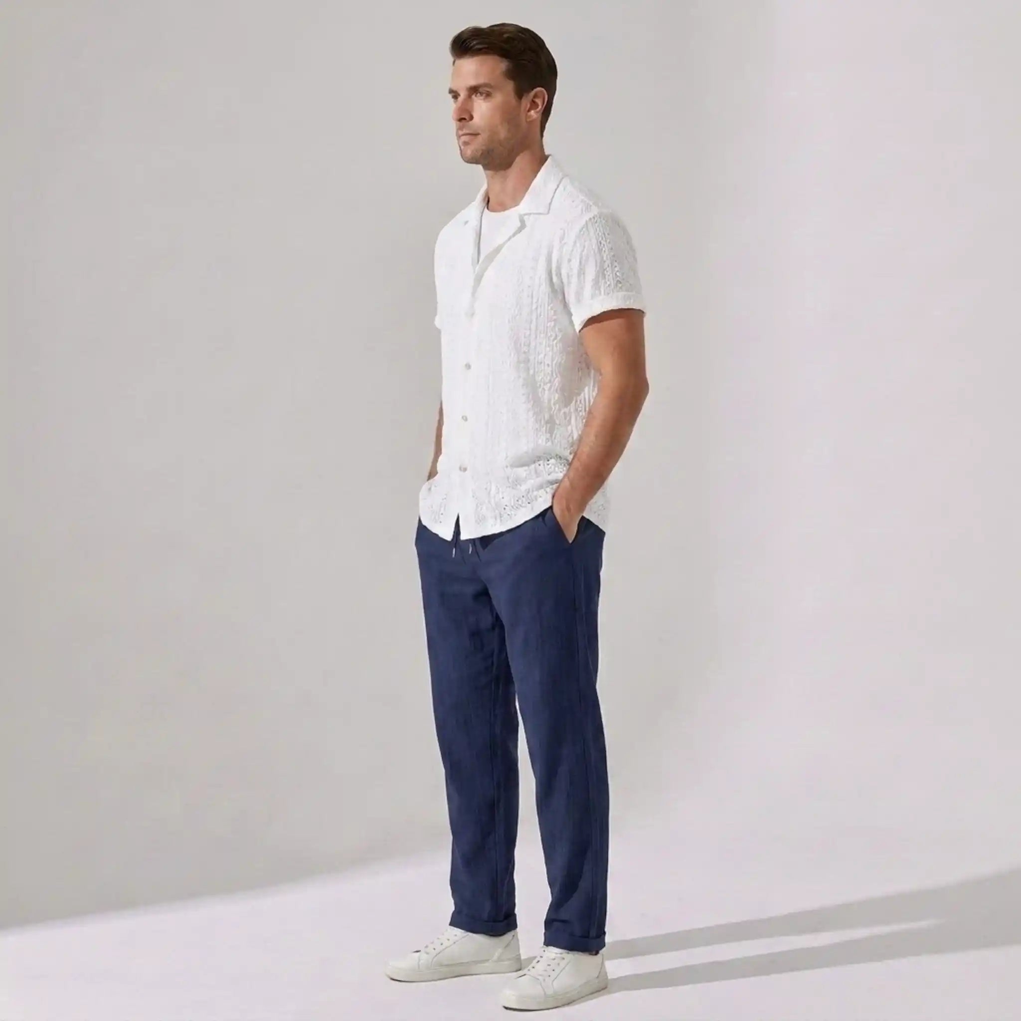 Man wearing a white patterned shirt, navy pants, and white shoes against a plain background