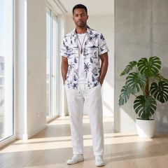 Man wearing a white shirt with blue palm tree pattern and white pants standing in a bright room with large windows and a plant.