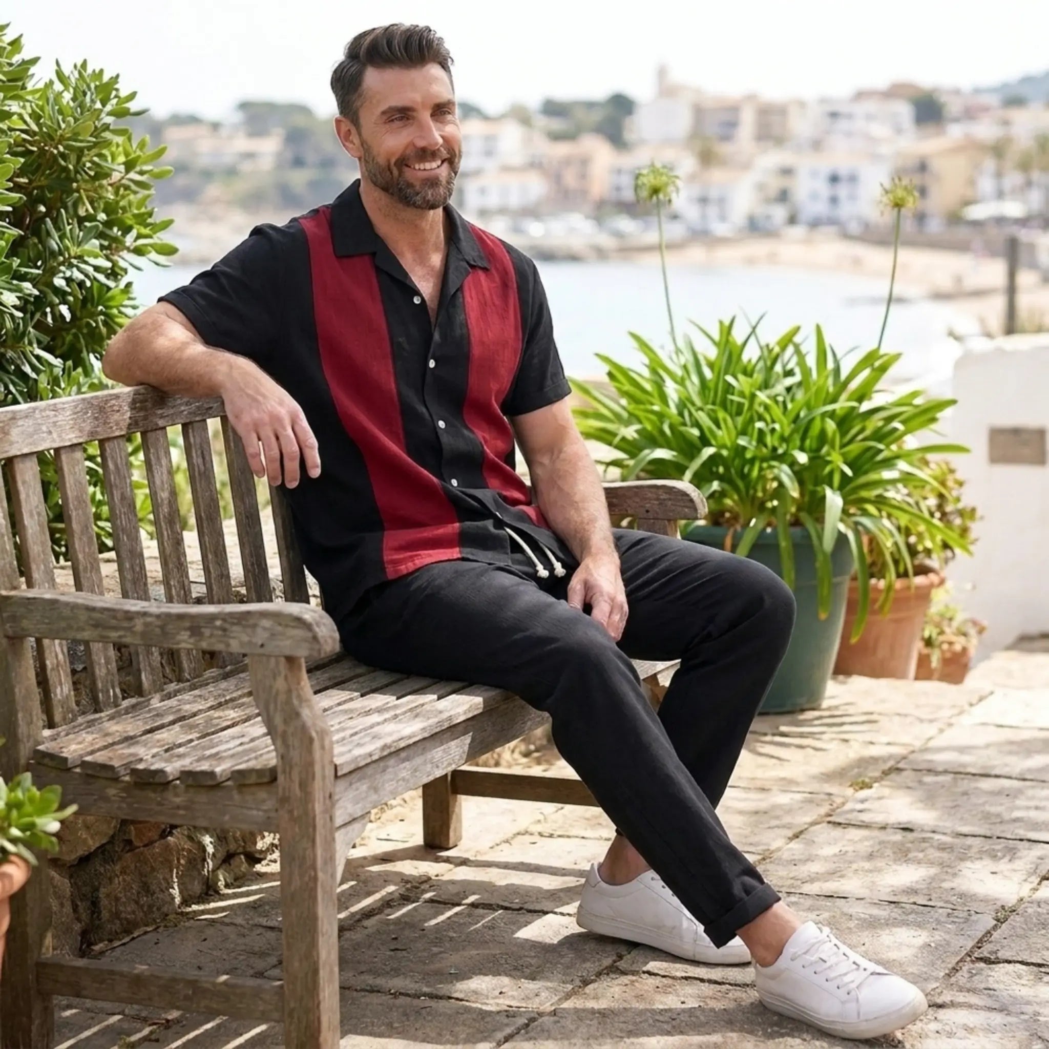 Man wearing black and red strip shirt and black trousers sitting on a wooden bench outdoors with a scenic background