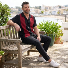 Man wearing black and red strip shirt and black trousers sitting on a wooden bench outdoors with a scenic background