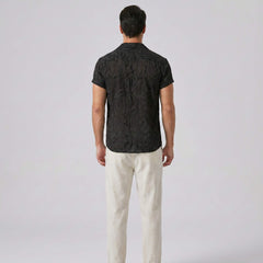 Man wearing a black patterned shirt and white pants on a light gray background