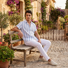Man wearing light blue and white stripe shirt and white pants sitting on a wooden bench in an outdoor setting with flowers and buildings in the background