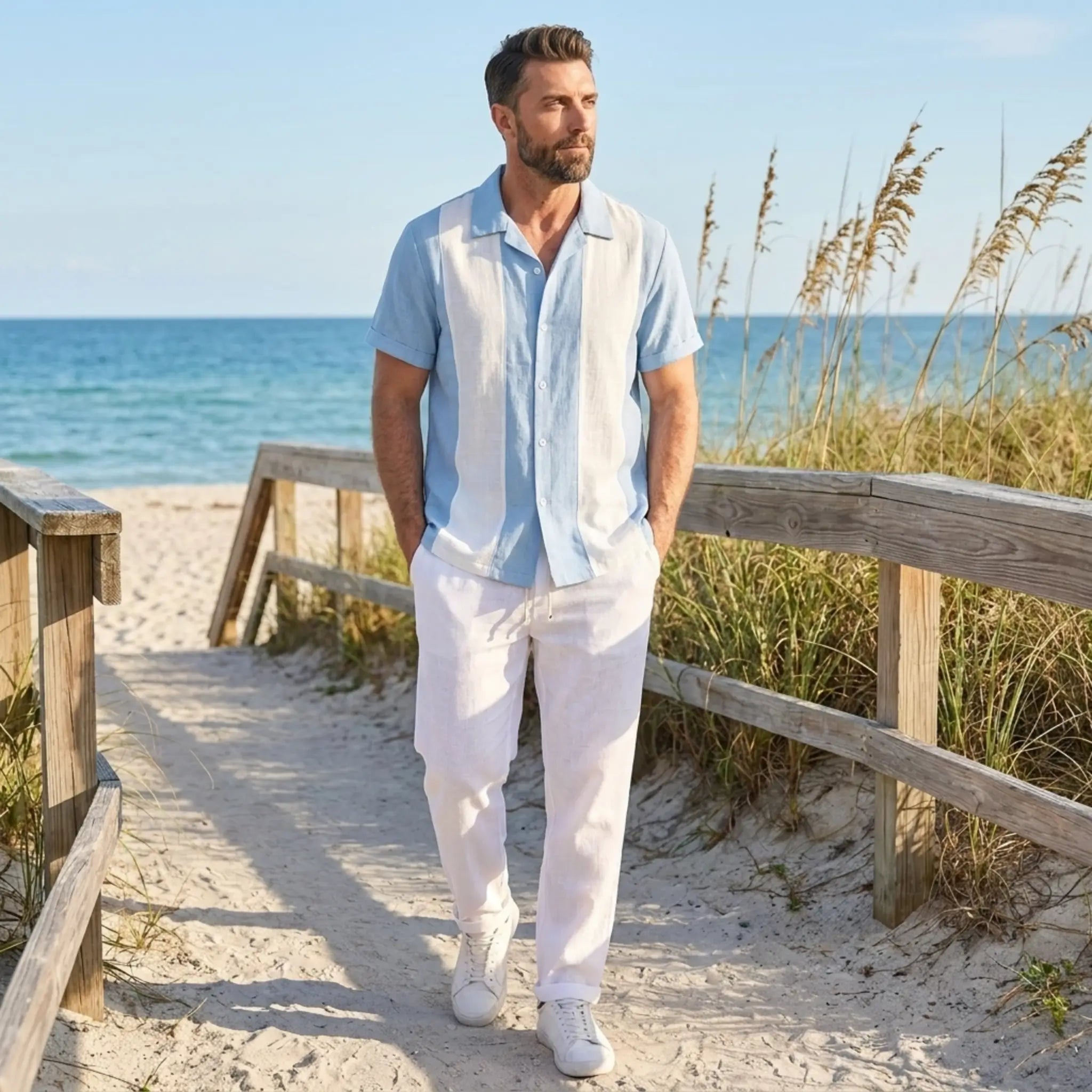 Man with blue polo and white trouser walking on a sandy path by the ocean with grasses and wooden railings.