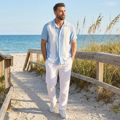 Man with blue polo and white trouser walking on a sandy path by the ocean with grasses and wooden railings.