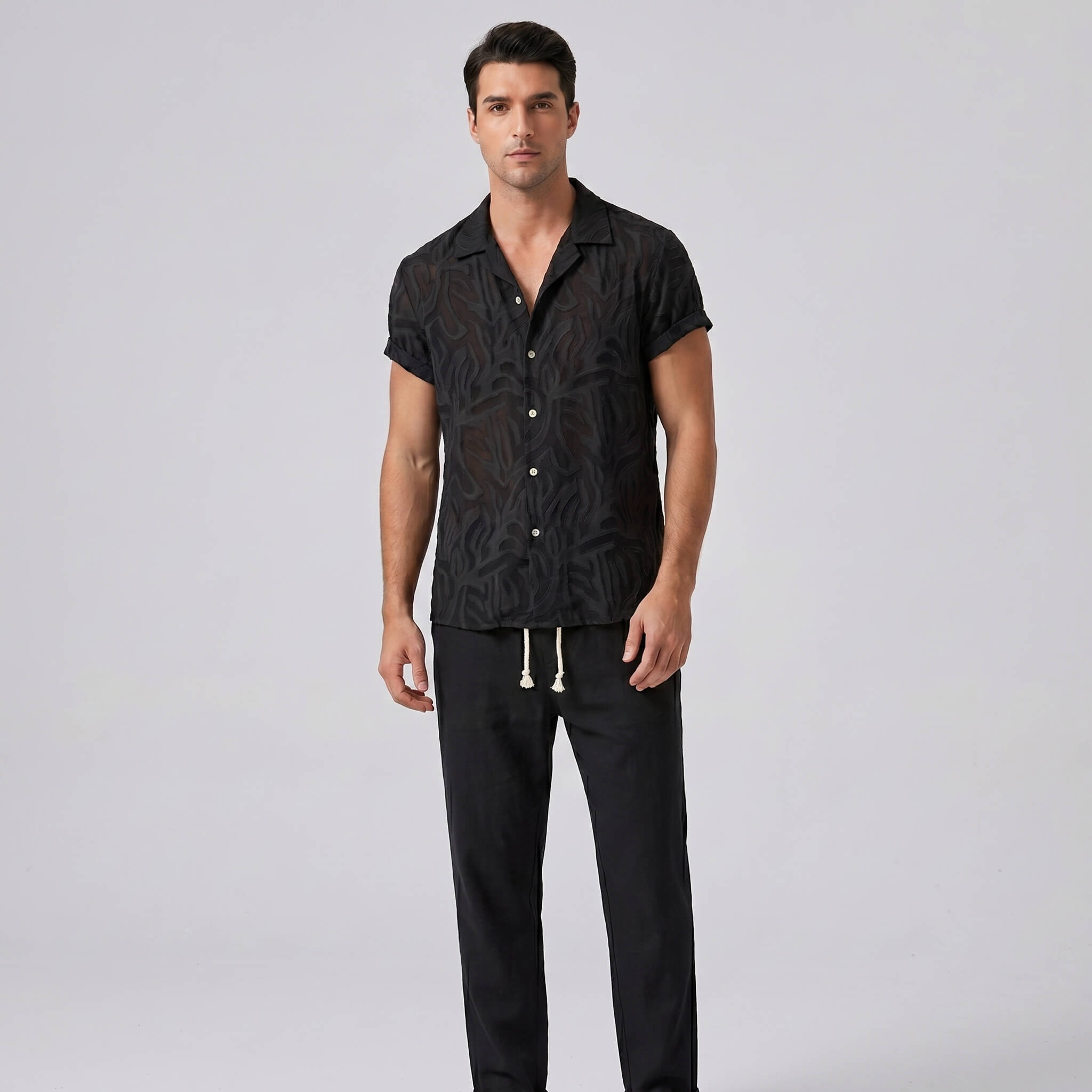 Man wearing a black patterned shirt and black pants on a light gray background