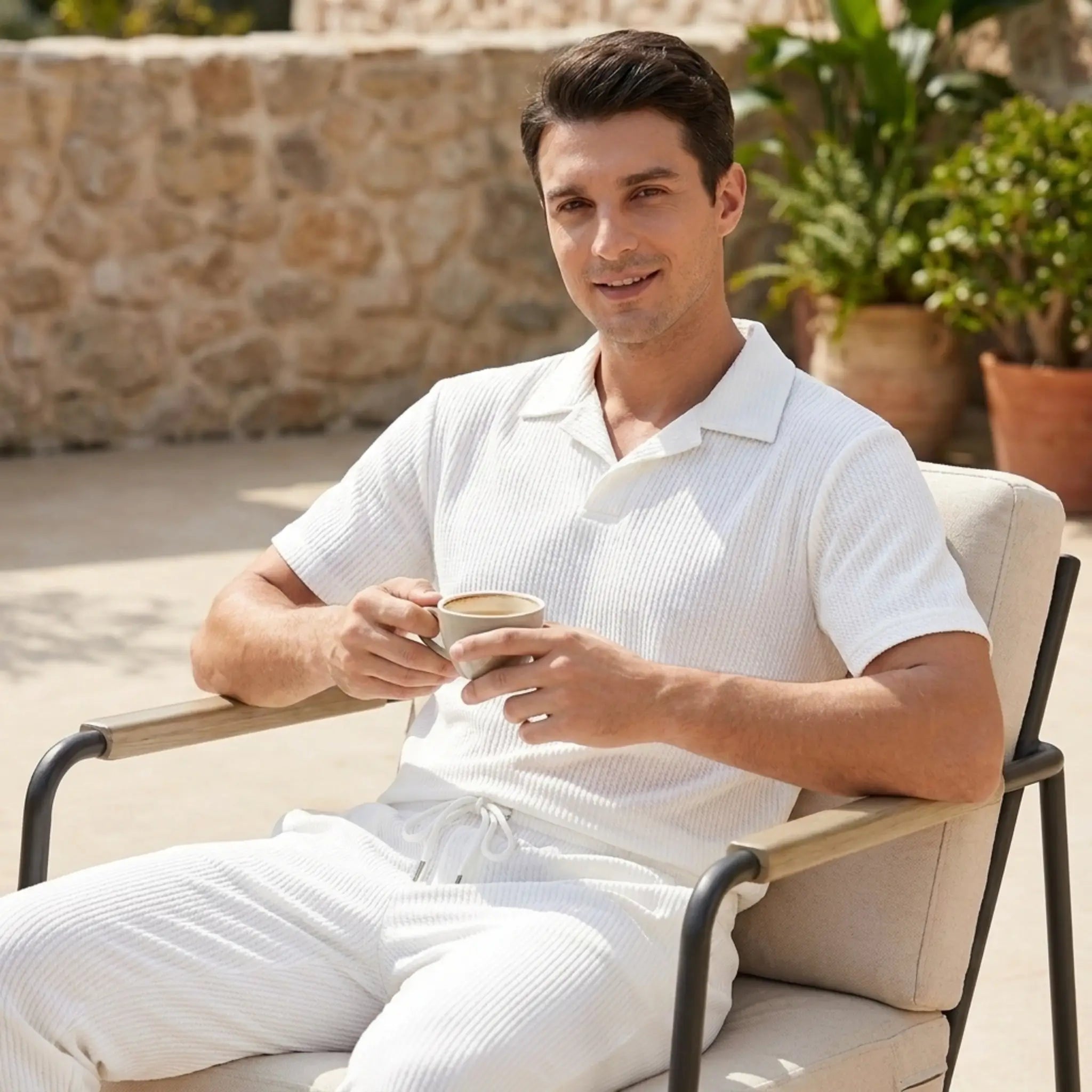 man wearing white polo and pants holding a cup of coffee while sitting on a sofa