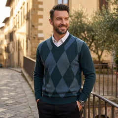 A smiling man wearing a dark teal and slate green argyle sweater over a white collared shirt, standing on a sunlit cobblestone street with buildings and a railing in the background.