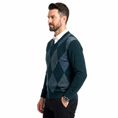 A man wearing a dark teal and green argyle sweater over a white collared shirt and black trousers, standing against a plain white background.