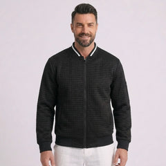 Man wearing MannerCo black quilted zip-up baseball jacket with stand collar, front view on plain background