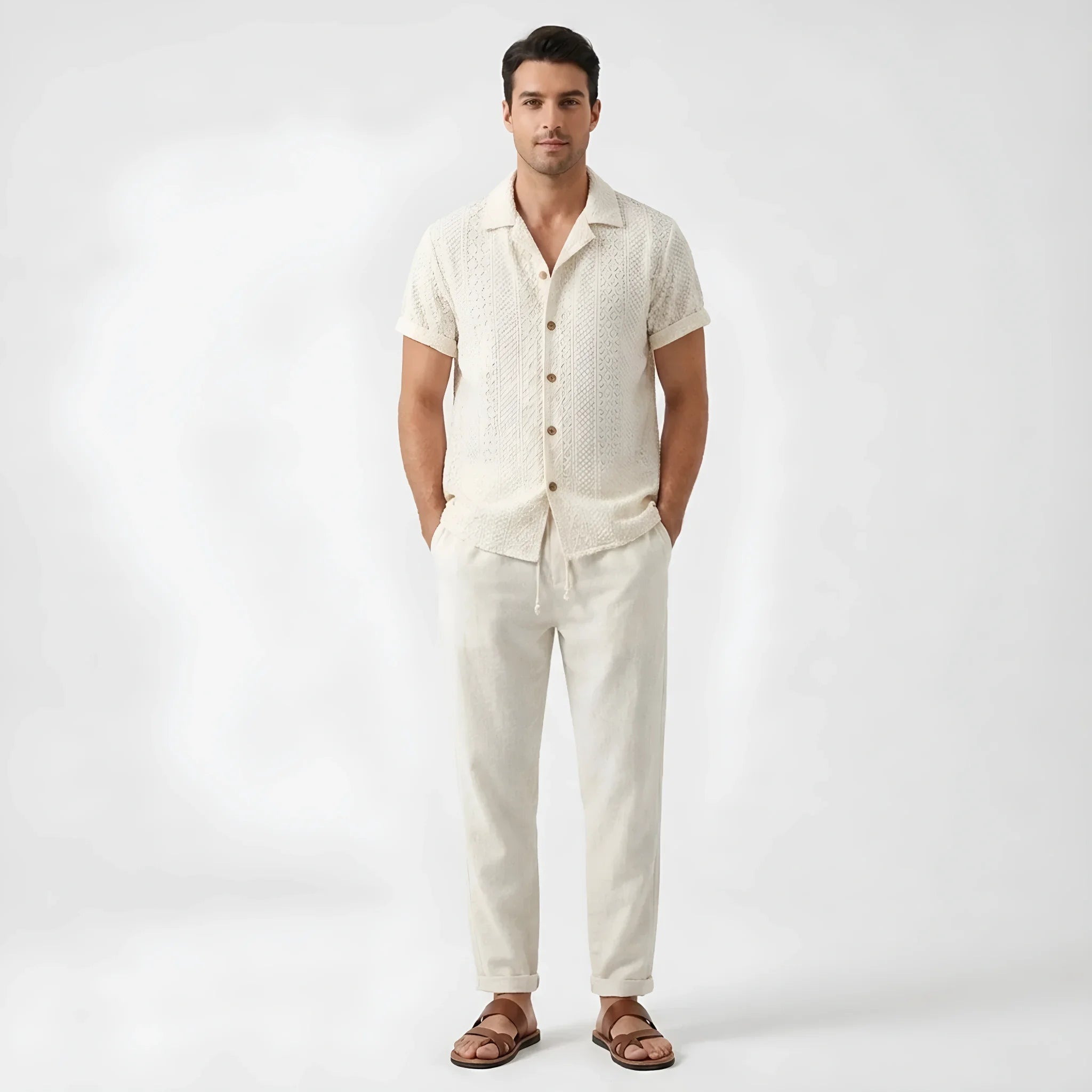 Man wearing a beige floral checkerboard jacquard Cuban shirt and straight leg linen pants front view on white background