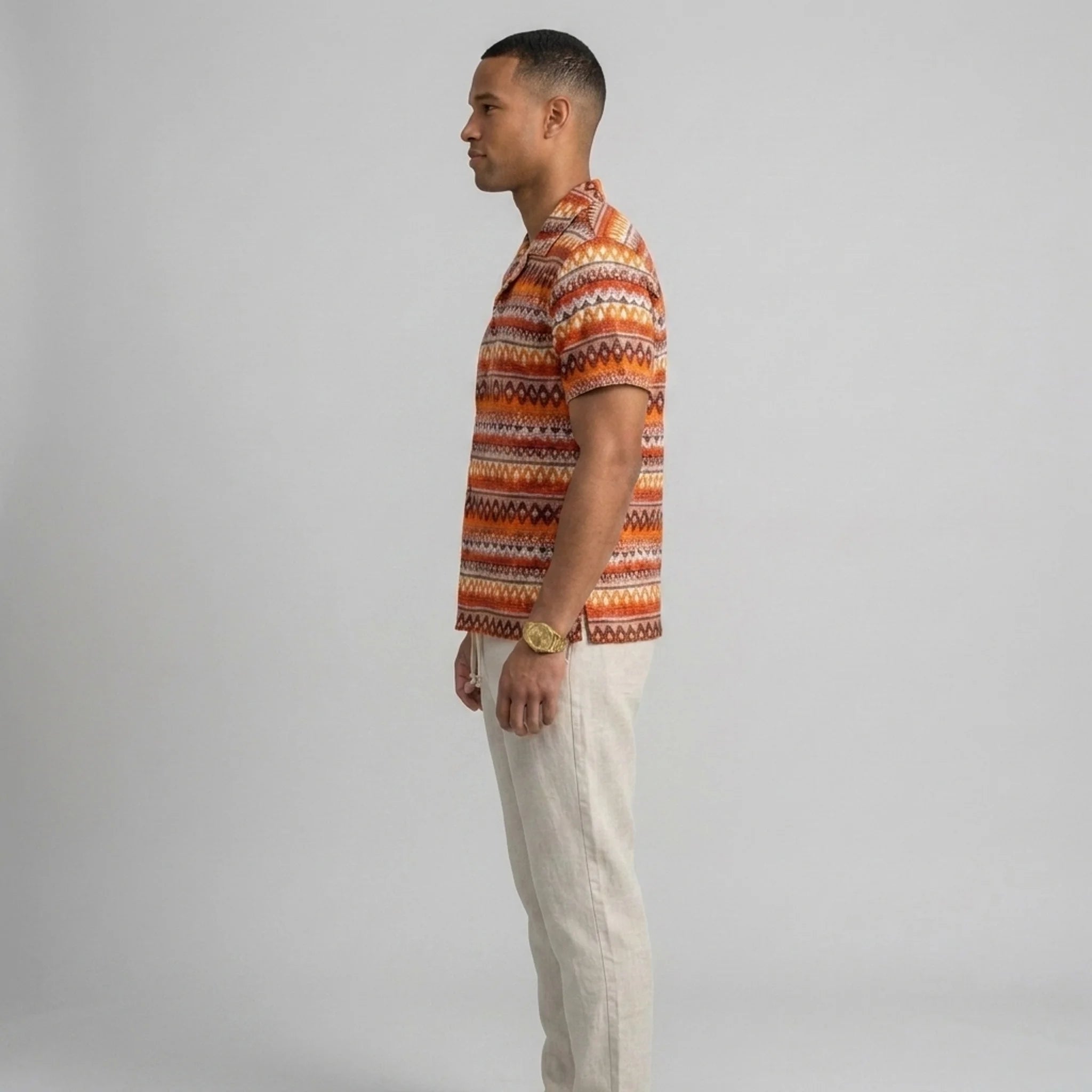 Side view of man in beige geometric Cuban shirt and straight leg linen pants on plain background