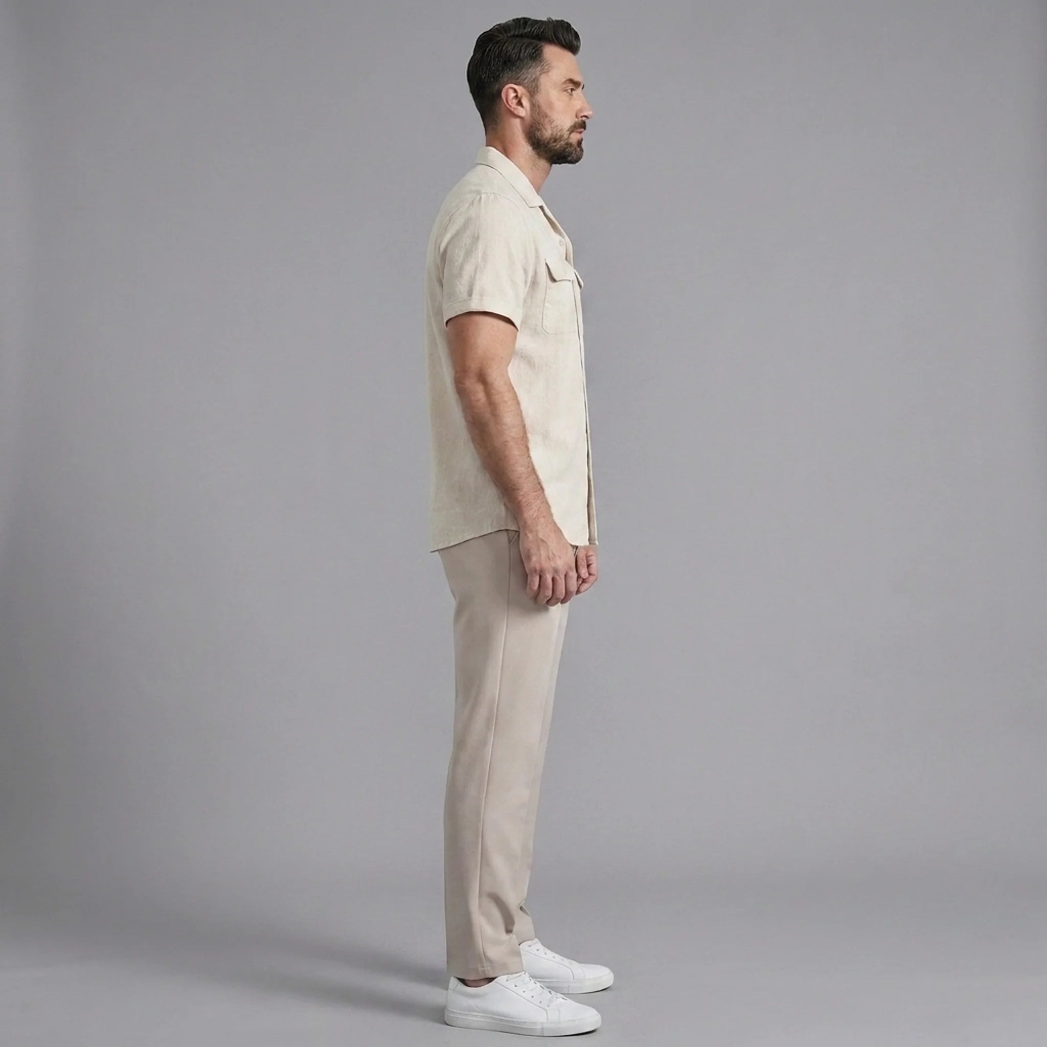 Side view of man in beige linen Cuban collar shirt and slim fit tapered trousers on plain background