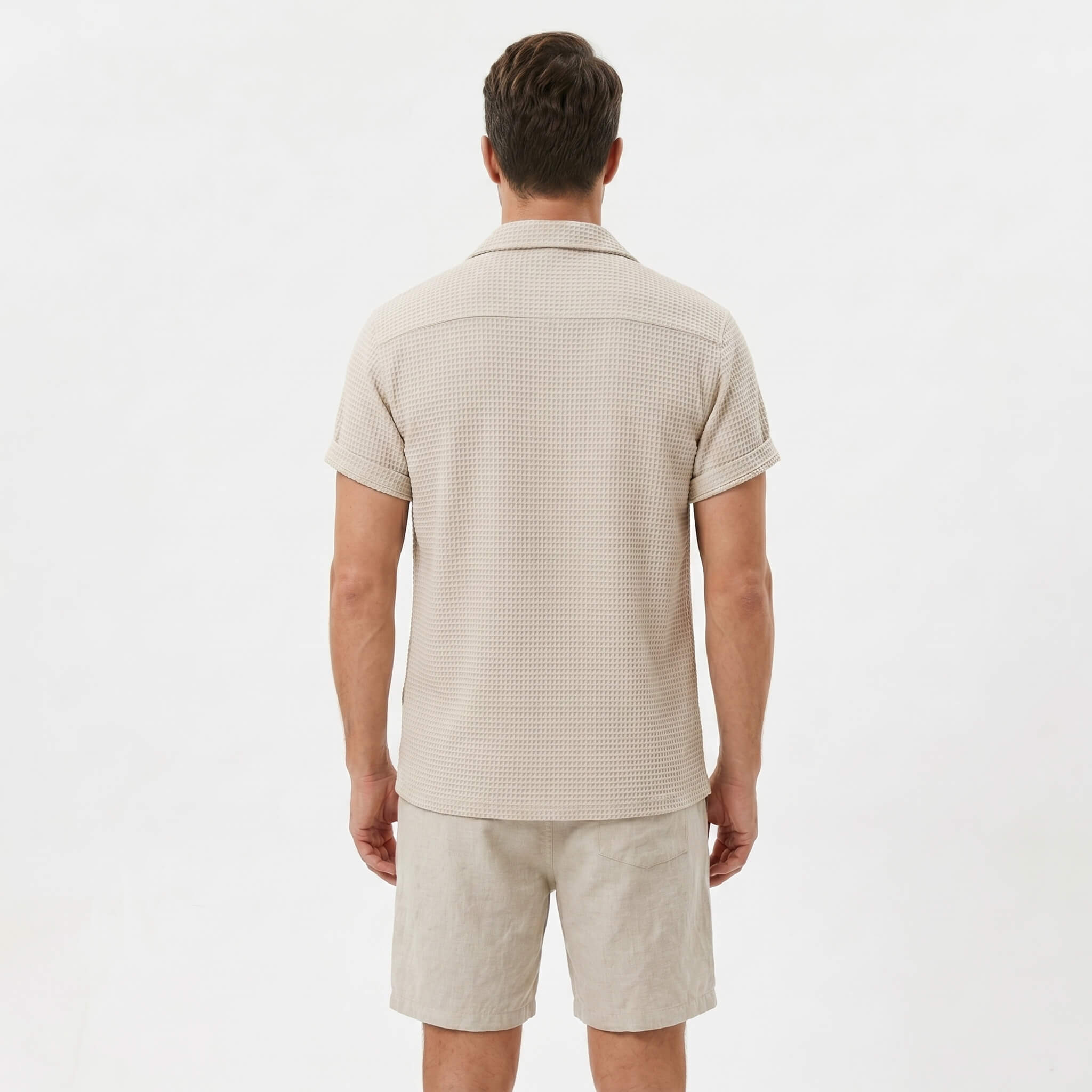 Side view of man in beige textured Cuban shirt and linen cotton drawstring shorts on plain background