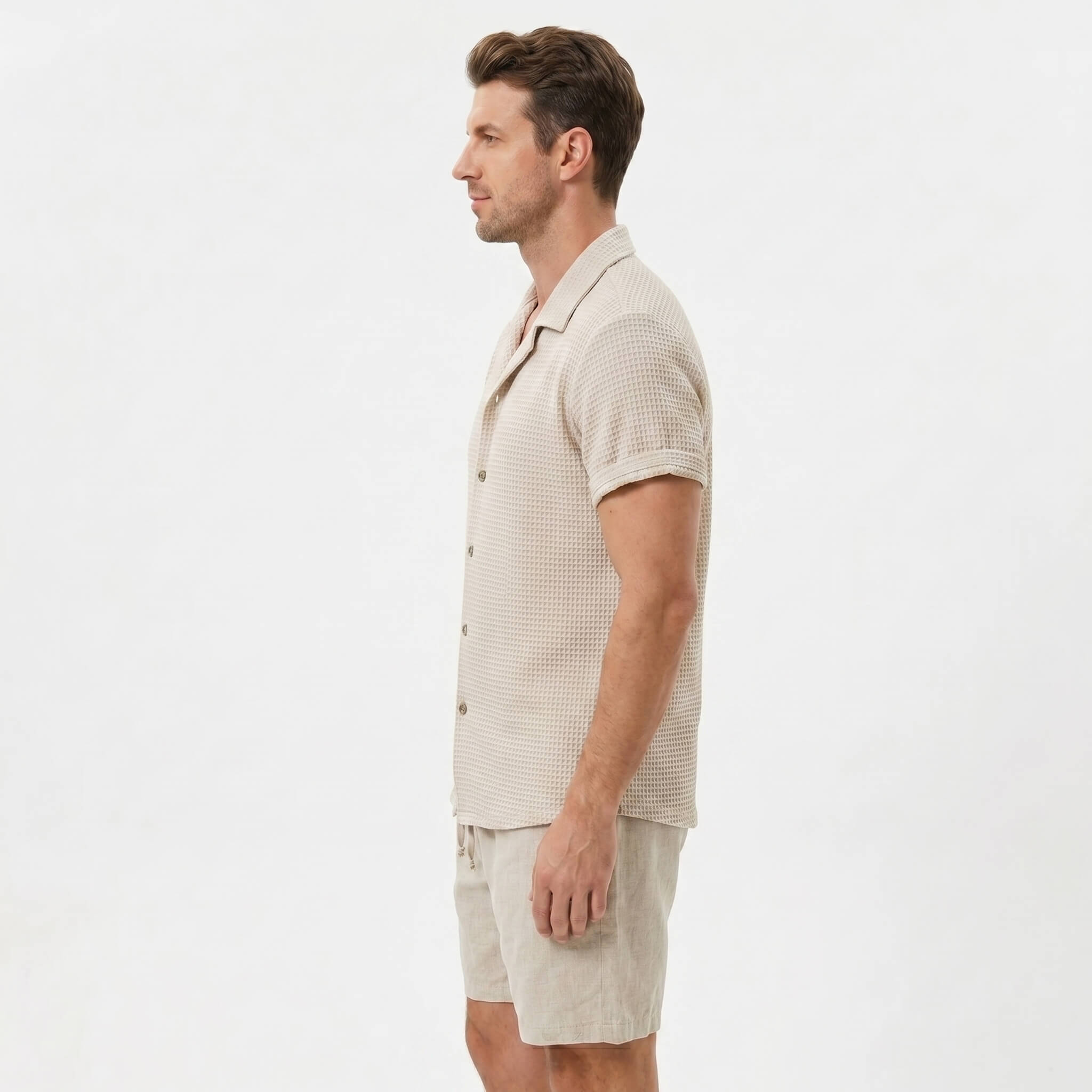 Three quarter view of man in beige textured Cuban shirt and linen drawstring shorts on plain background