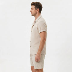 Three quarter view of man in beige textured Cuban shirt and linen drawstring shorts on plain background