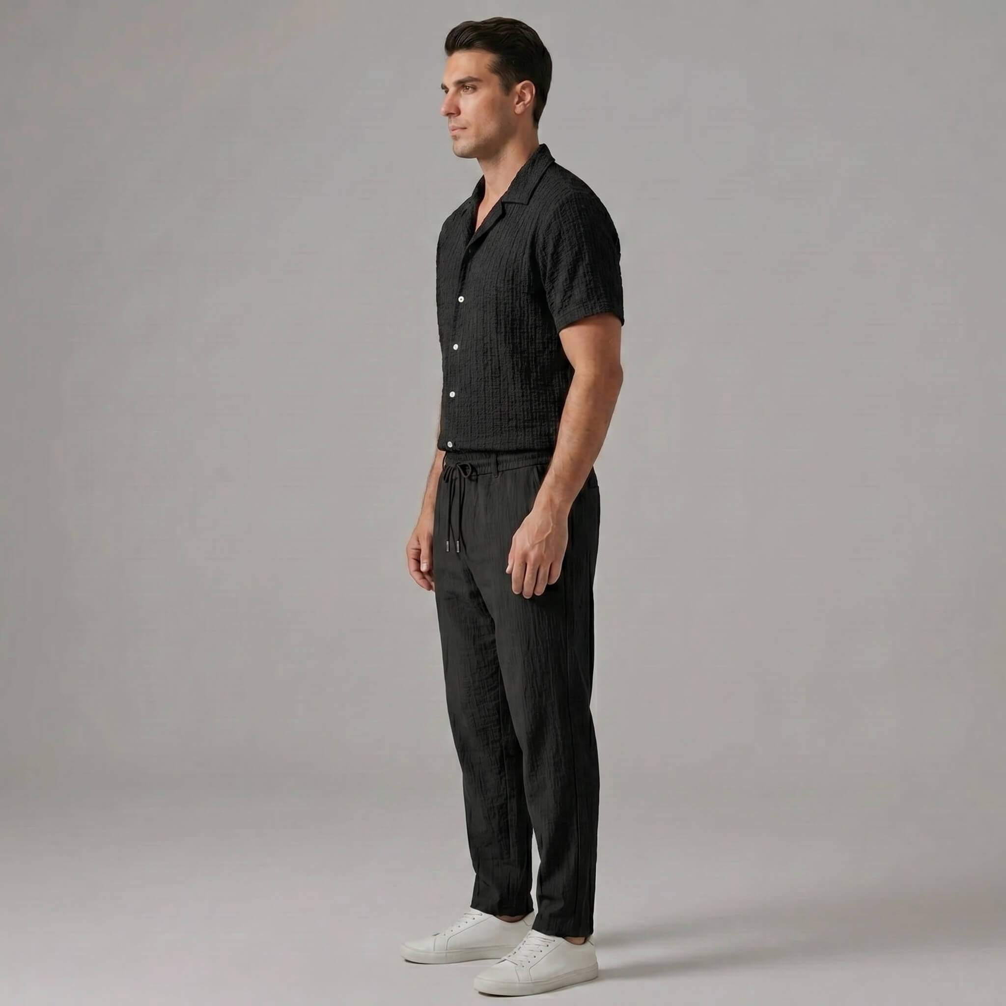 Side view of man in black jacquard Cuban shirt and crinkle straight leg pants on plain background