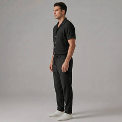 Side view of man in black jacquard Cuban shirt and crinkle straight leg pants on plain background