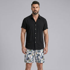 Man in black linen rayon shirt and white palm tree swim shorts walking in sunny outdoor scene