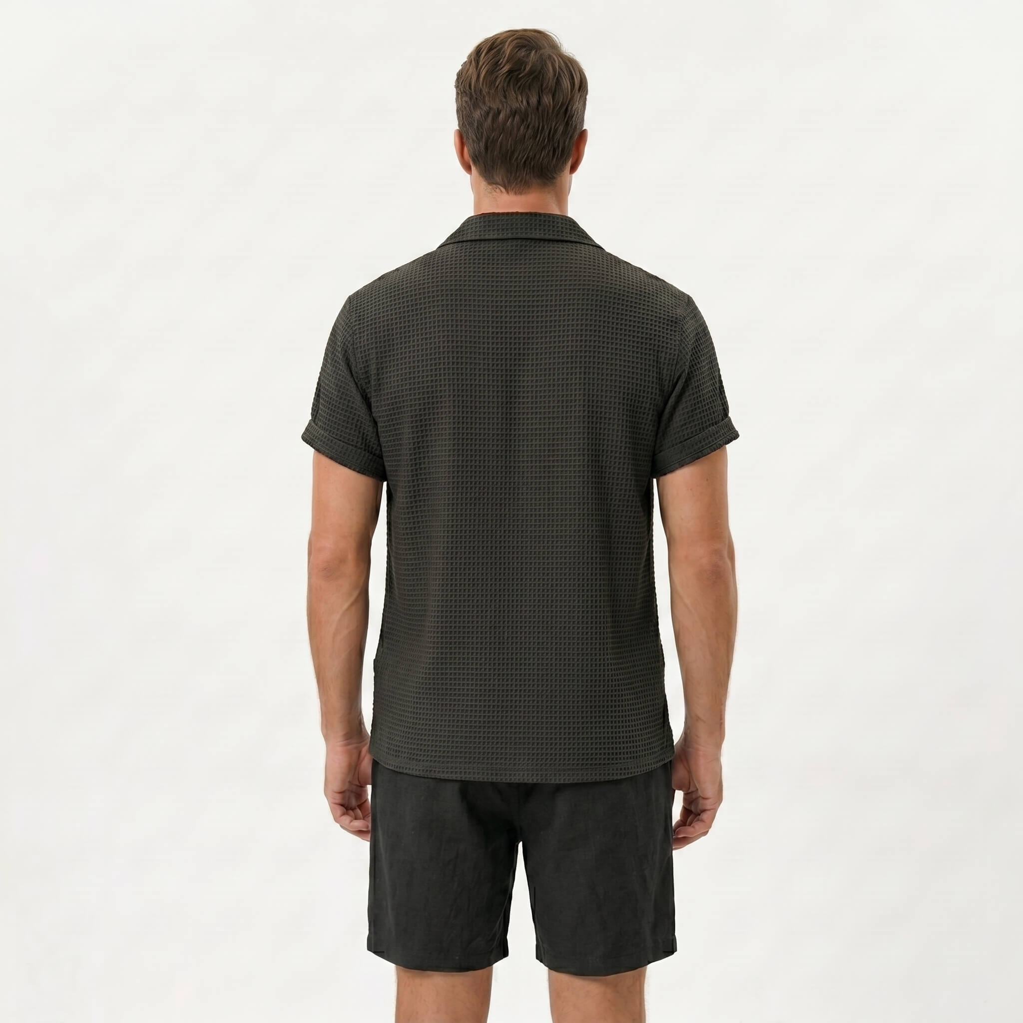 Side view of man in coffee brown textured Cuban shirt and linen drawstring shorts on plain background