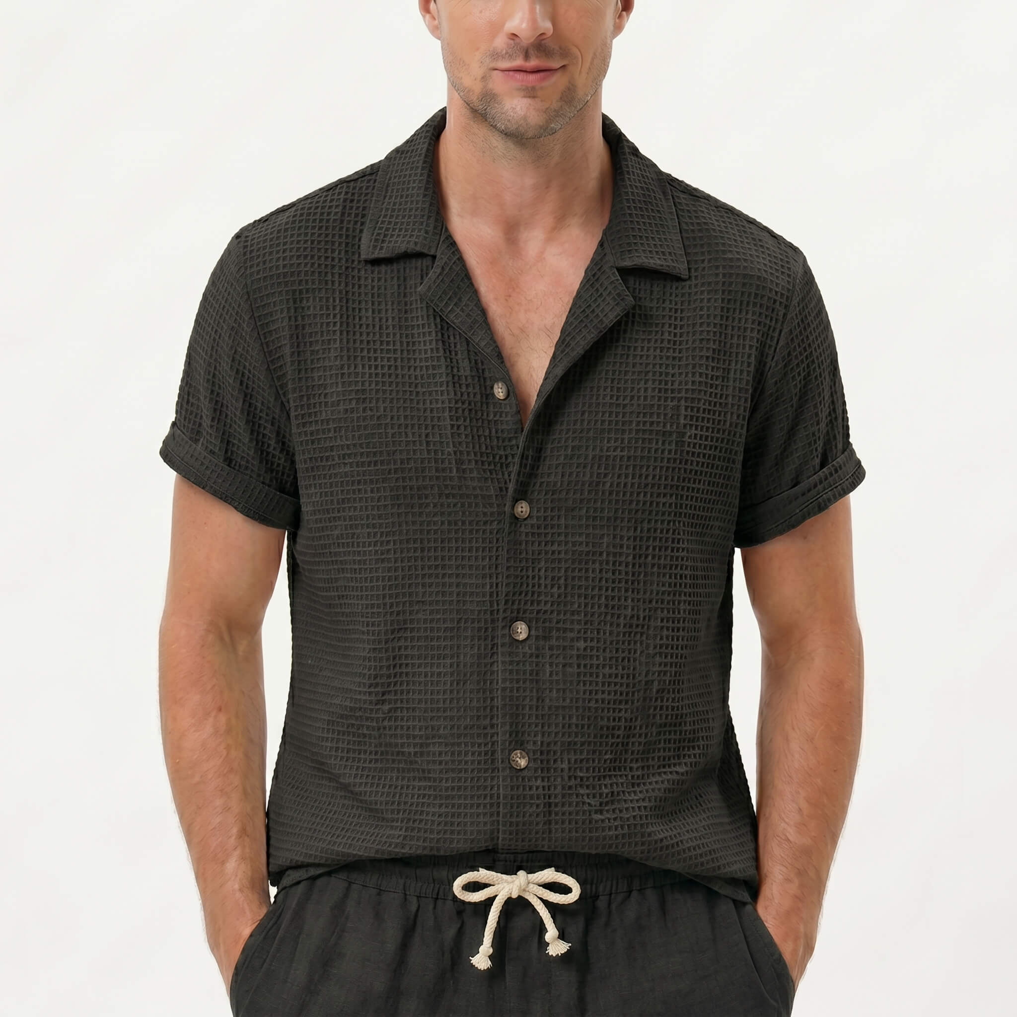 Three quarter view of man in coffee textured Cuban shirt and linen drawstring shorts on plain background