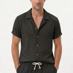 Three quarter view of man in coffee textured Cuban shirt and linen drawstring shorts on plain background