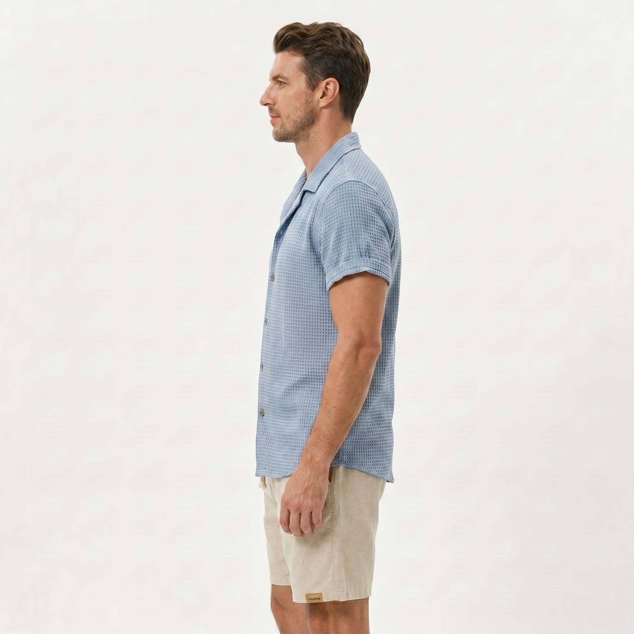 Side view of man in light blue textured Cuban shirt and linen drawstring shorts on plain background
